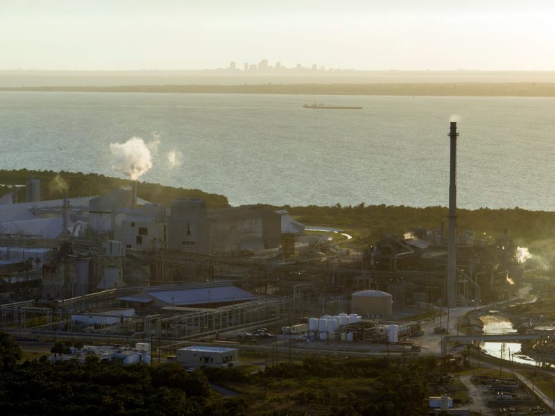 An aerial view of a large industrial complex featuring smokestacks emitting white steam, various storage tanks, and a tall chimney, situated along a coastline. Behind the facility lies a wide, calm body of water with a barge floating in the distance. On the far horizon, a city skyline is faintly silhouetted against a hazy, golden sky.