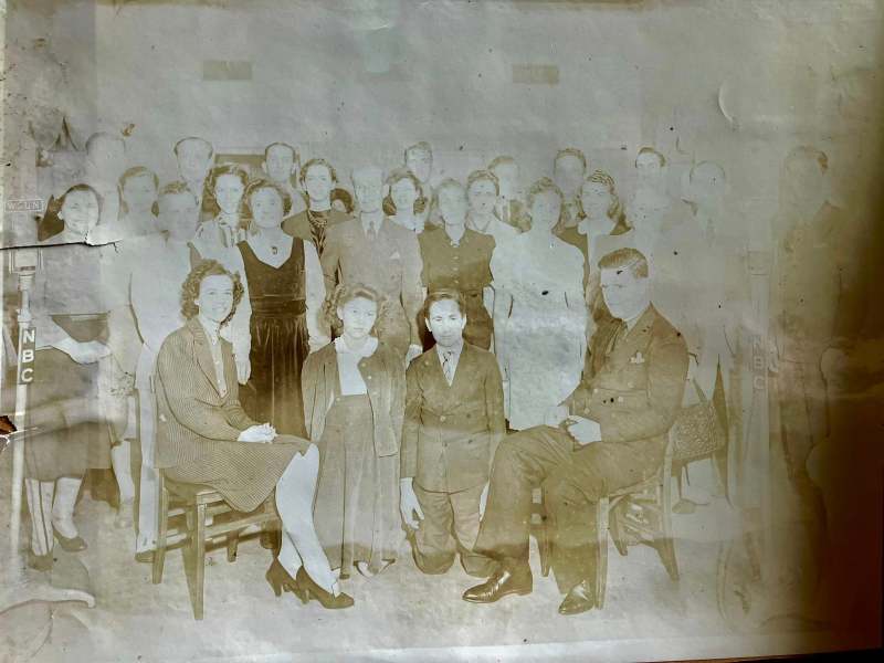A heavily faded, sepia-toned vintage black-and-white photograph of a large cast or studio group from the mid-20th century. The subjects are formally dressed, with some standing in rows and two seated figures in the foreground, and the image is marked by blemishes and water stains.