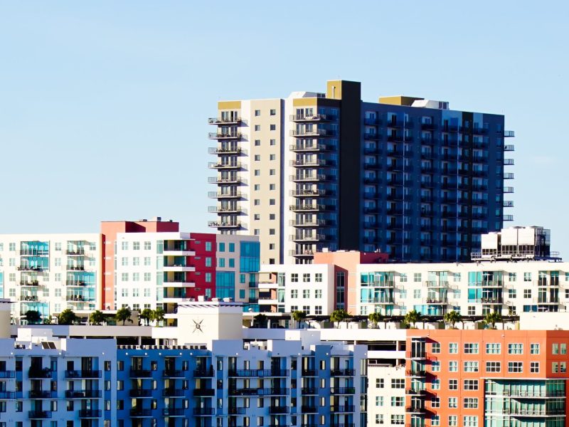 Modern apartment buildings in downtown Tampa, featuring a mix of high-rise and mid-rise structures. The buildings have colorful facades in shades of white, red, and blue, with many balconies and glass windows. A taller dark gray tower rises in the background against a clear blue sky.