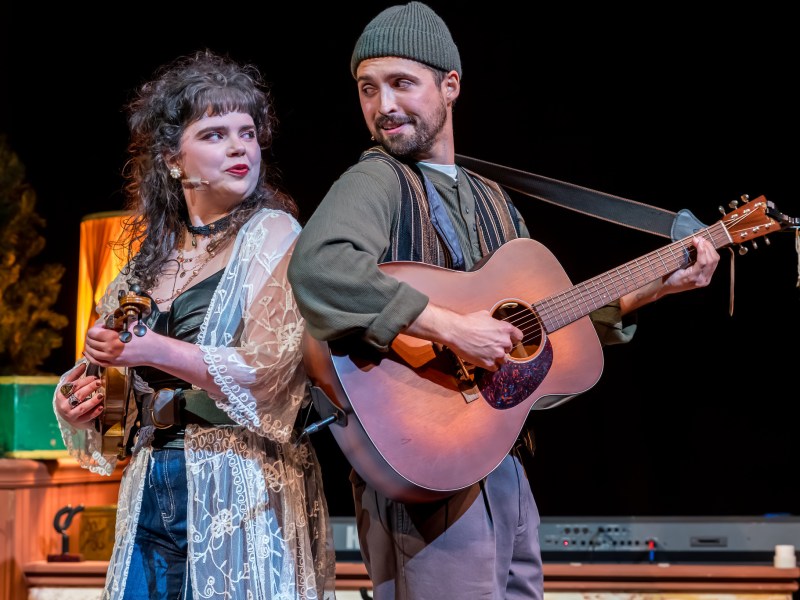 A close-up theatrical shot of two musicians. One person, wearing an earth-toned outfit, is singing and playing a large acoustic guitar, while the other person, in dark jeans and a sheer top, holds a small fiddle or violin and looks off to the side.
