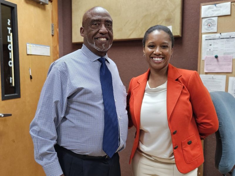 A portrait of a bald, dark-skinned man with a gray goatee, wearing a checked dress shirt and a navy tie, standing next to a smiling woman with dark hair pulled back. The woman is wearing a white blouse and a bright orange blazer. They are standing indoors next to a wood-paneled wall and a bulletin board.