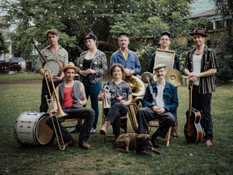 The eight members of the band Tuba Skinny pose in a grassy backyard setting with their instruments, including a banjo, tuba, trumpet, trombone, washboard, and guitars, with a brindle dog resting in the foreground.
