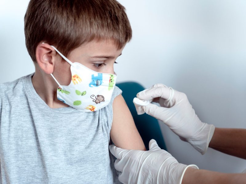 Close-up of a child wearing a face mask being prepared for a vaccination by a person wearing white medical gloves, who is swabbing the child's upper arm with cotton.