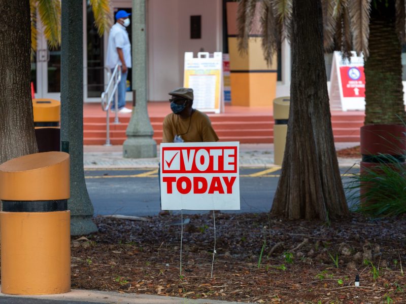 A temporary red and white election sign that reads "VOTE TODAY" is staked into the ground between two palm trees outside a polling place. A person wearing a mask is partially visible behind the sign.