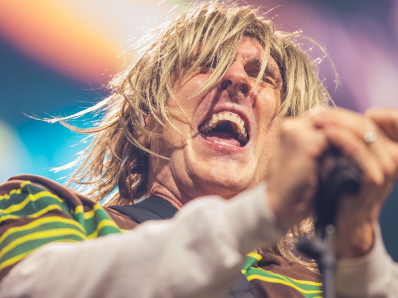 A close-up of a person with long, shaggy blonde hair singing passionately into a microphone. Their mouth is wide open and their eyes are squinted as they tilt their head back in a moment of performance. They are holding the microphone with both hands.