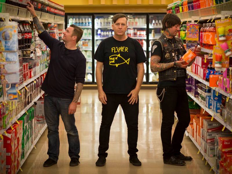 Three band members pose in the middle of a grocery store. The subject on the left is reaching for a beverage, and the person on the right is holding an orange juice container, both wearing denim. All three subjects have visible tattoos on their arms.