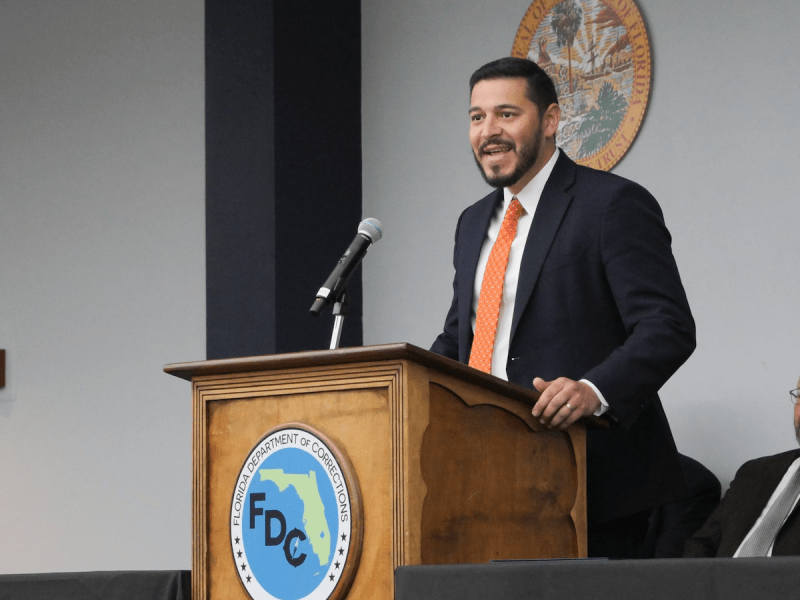 A smiling, dark-haired man in a navy suit, white shirt, and bright orange tie speaks enthusiastically into a microphone from a wooden podium. The Florida Department of Commerce (FDC) logo is visible on the podium's front, and the Great Seal of the State of Florida is on the wall behind him.