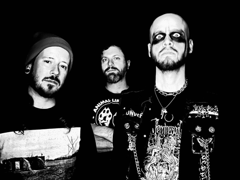 A trio of serious-looking musicians. The subject on the left wears a beanie and large ear gauges. The central person wears a dark t-shirt, and the person on the right wears a patched, chain-adorned vest and metal necklaces.