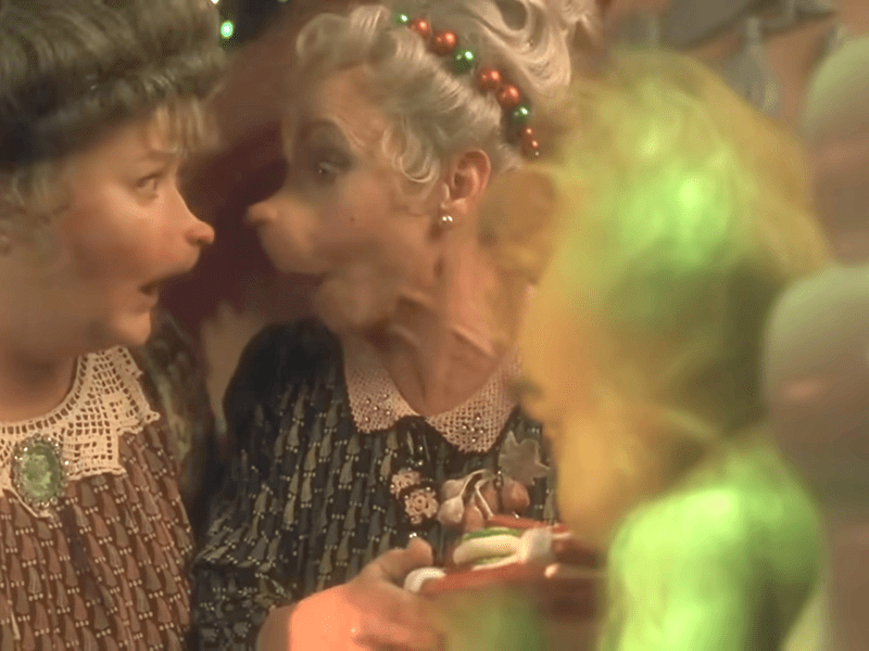 A shot from the movie "How the Grinch Stole Christmas:" Two female Who characters with distinctive facial prosthetics and elaborate hairstyles look at one another with expressions of shock or surprise. The woman on the left wears a brown dress with a white lace collar and a green brooch, while the woman on the right wears a dark patterned dress and a festive red and green beaded headband. In the foreground, the blurry green fur of the Grinch is partially visible.