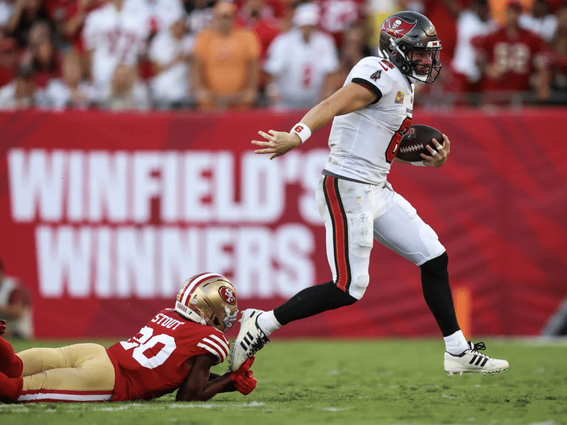 A Tampa Bay Buccaneers quarterback in a white uniform runs with the football, successfully evading a low tackle attempt by a San Francisco 49ers defender.