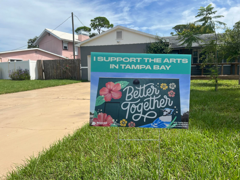 A yard sign on green grass in front of a driveway and house. The sign has a teal border and text at the top that reads, “I SUPPORT THE ARTS IN TAMPA BAY.” Below is an image of a mural with colorful flowers, a blue jay, and large white script that says “Better Together.”
