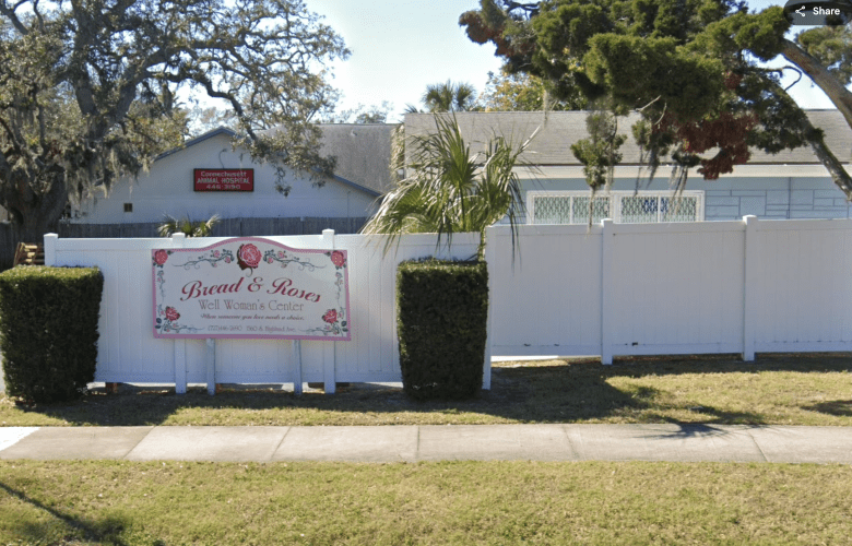 A daytime street view of a small community building behind a white fence and trimmed hedges. The sign is visible in the foreground, with the single-story clinic building and surrounding residential trees in the background.
