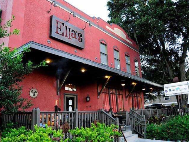 A low-angle, exterior shot of a two-story restaurant named "Ella's." The building is painted bright red with black trim, and a black metal sign with "Ella's" in a distinctive font juts out from the wall. A dark awning covers a front patio area, which is accessed by stairs and enclosed by a rustic, dark-colored fence. Lush green foliage and trees surround the entrance, and a lit torch-style lamp is visible near the stairs.