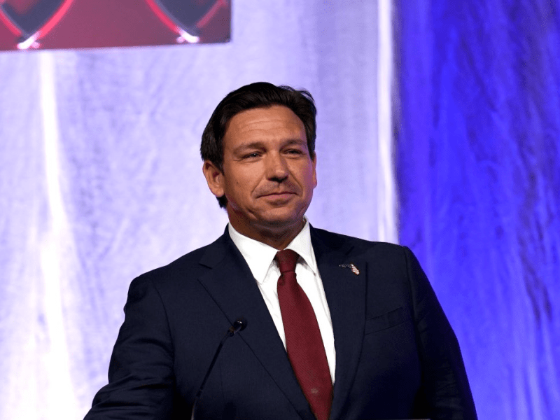 A political or business speaker with short, dark hair, wearing a suit jacket with an American flag pin on the lapel. The background is composed of dark red and blue draping, illuminated by bright stage lights.