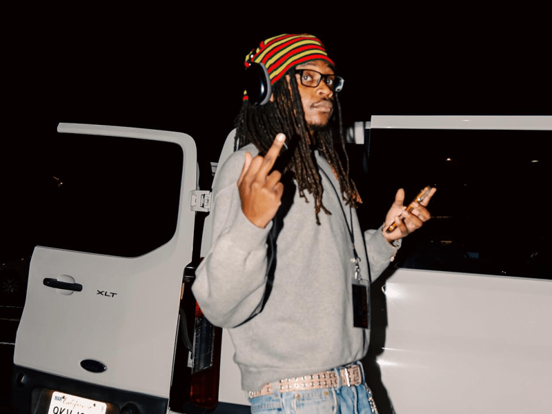 An individual with long dreadlocks and glasses, wearing a striped beanie and a grey sweatshirt, stands in front of an open white Ford XLT van at night. They are holding a smartphone in one hand and gesturing toward the camera.