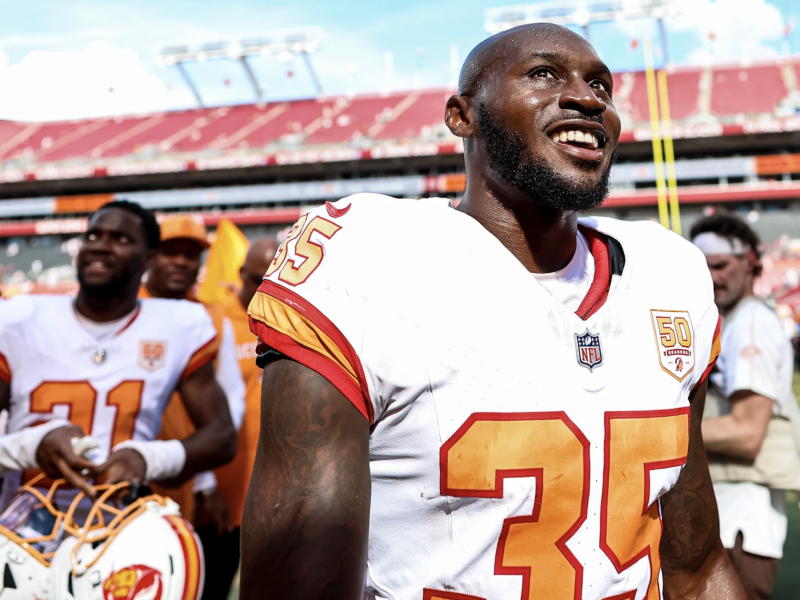 Jamel Dean of the Tampa Bay Buccaneers, wearing a white No. 35 jersey with red and gold trim, smiles as he walks off the field after a game at Raymond James Stadium. Teammates, including one holding a helmet, and staff members are gathered in the background under the bright afternoon sun.