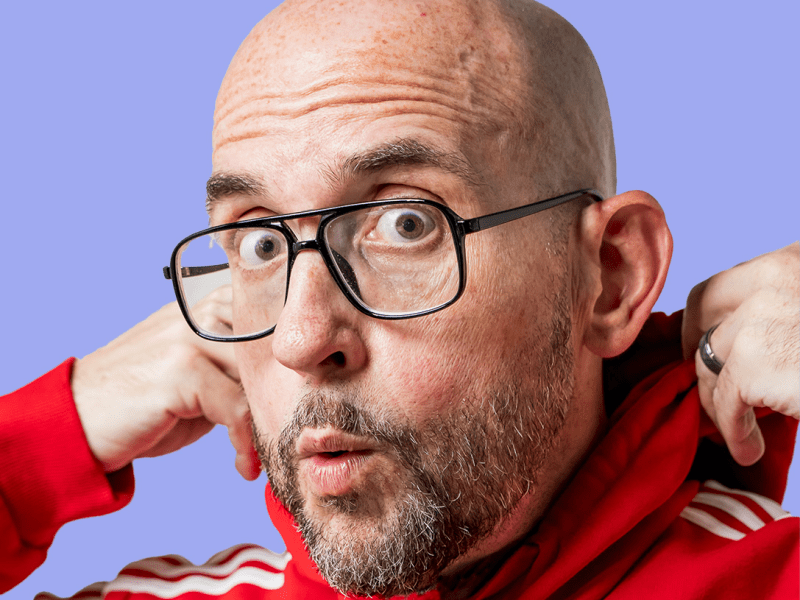 Close-up portrait of comedian Johnny B wearing black-rimmed glasses and a red Adidas hoodie, making a playful surprised face while tugging on his collar, set against a purple background.