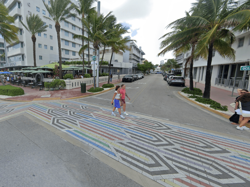 Street view of Ocean Drive in Miami Beach showing a colorful rainbow-pattern crosswalk with geometric designs. Palm trees line the street alongside art deco-style hotels and restaurants with outdoor seating, including Hotel Victor. Several pedestrians cross the street while others stand near the corner under a green “Ocean Dr” street sign. Cars are parked along the road in the background.