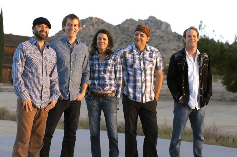 A group portrait of the five members of the band Donna the Buffalo standing outdoors in a desert landscape. From left to right: Kyle Spark, Mark Raudabaugh, Tara Nevins, Jeb Puryear, and David McCracken. They are all smiling and dressed in casual, roots-rock attire, including flannel shirts and denim. Tara Nevins, in the center, wears a blue plaid shirt and a decorative belt, while Jeb Puryear to her right wears a patterned hat. The background features a large, rocky hill under the soft glow of golden hour light, with a rustic wooden building visible on the left.