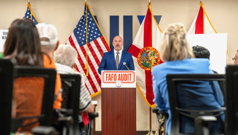 A bald man in a blue suit stands at a podium speaking into a microphone during a formal event. The podium features a prominent orange sign that reads "FAFO AUDIT." Behind the speaker, American and Florida state flags are displayed, while the backs of seated audience members frame the foreground.