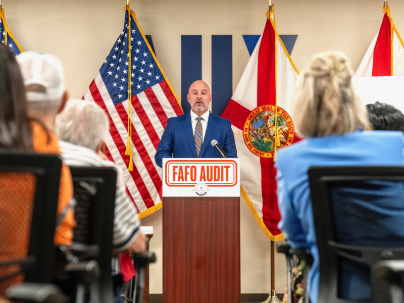 A bald man in a blue suit stands at a podium speaking into a microphone during a formal event. The podium features a prominent orange sign that reads "FAFO AUDIT." Behind the speaker, American and Florida state flags are displayed, while the backs of seated audience members frame the foreground.
