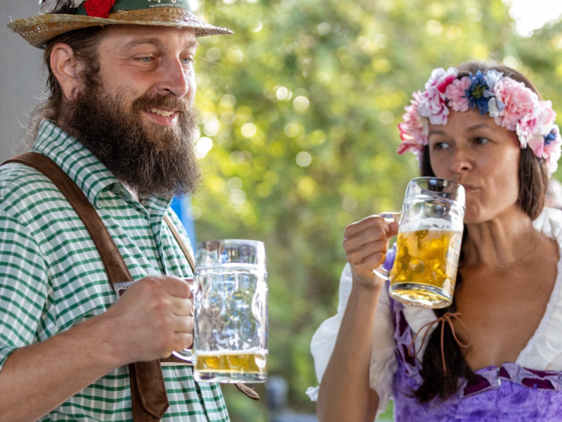 A man with a full beard and a straw hat with a feather and a red rose is smiling while holding a beer stein. He is wearing a green and white checkered shirt with brown suspenders. Beside him, a woman with a floral wreath on her head sips from a matching beer stein. She is wearing a purple and white traditional German dirndl with a white blouse. The background is a blurry outdoor setting.