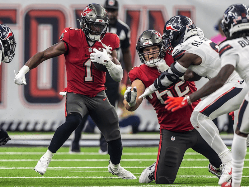 Tampa Bay Buccaneers running back Rachaad White (#1) carries the football while charging through defenders in a game against the Houston Texans. He wears a red jersey and black pants, with teammates blocking ahead as Texans players in white jerseys move in to tackle.