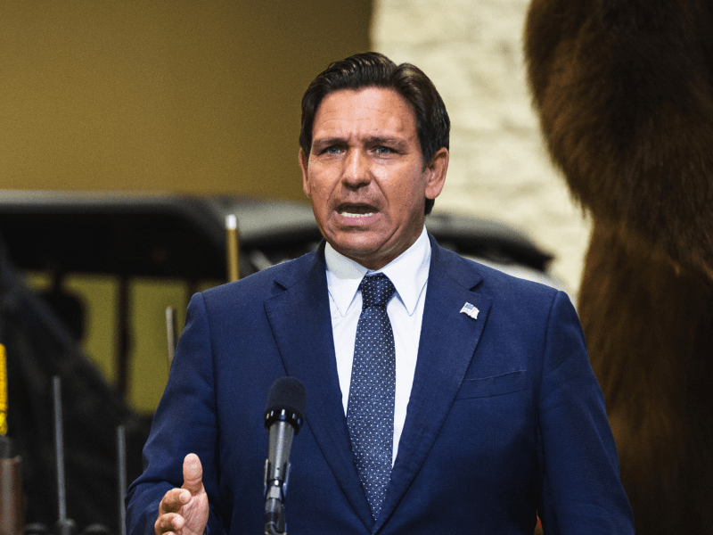 Florida Governor Ron DeSantis speaking at a press conference. He is wearing a dark blue suit with a patterned blue tie and a white shirt. An American flag pin is on his lapel. He has short, dark brown hair and is gesturing with his right hand as he speaks into a microphone. In the blurred background, there's a golf cart and what appears to be a large, taxidermied animal. The image is credited to Dave Decker.