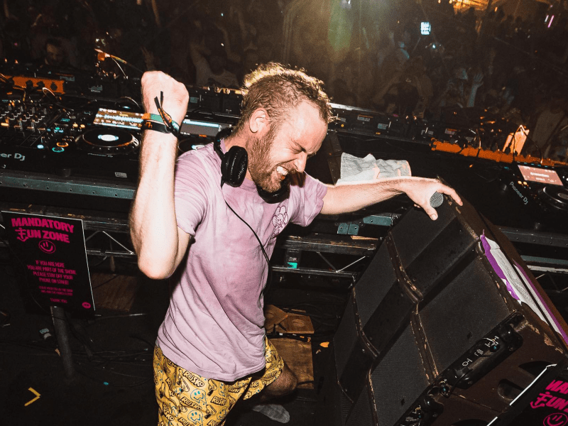 A musician with a short beard and headphones is performing at a music festival or concert. DJ equipment (Pioneer CDJs) is visible on the tables behind them, and warning signs reading "MANDATORY FUN ZONE" are visible on the speaker and surrounding area.