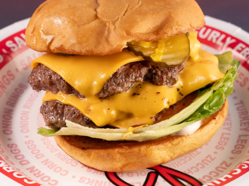 A juicy double cheeseburger served on a disposable white paper plate printed with red text like "ONION RINGS" and "CORN DOGS." The burger features two beef patties, melted yellow cheese, pickles, and lettuce on a soft, toasted bun.