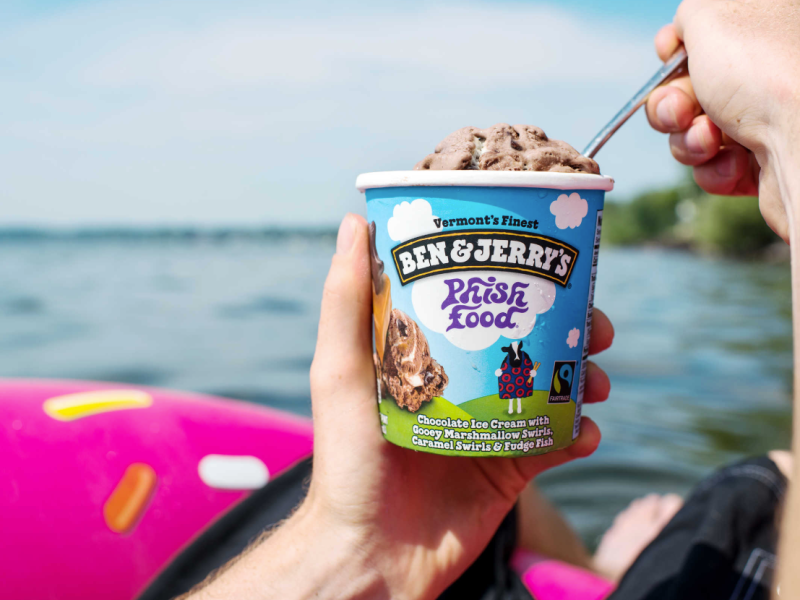 A first-person perspective shows a hand holding a pint of Ben & Jerry's "Phish Food" ice cream against a sunny backdrop of a blue lake. Another hand is using a spoon to scoop the chocolate ice cream. The blue pint container features the brand's logo and cloud imagery. A bright pink inflatable pool float with yellow sprinkles is partially visible in the lower left corner, suggesting a summer boating or swimming setting.