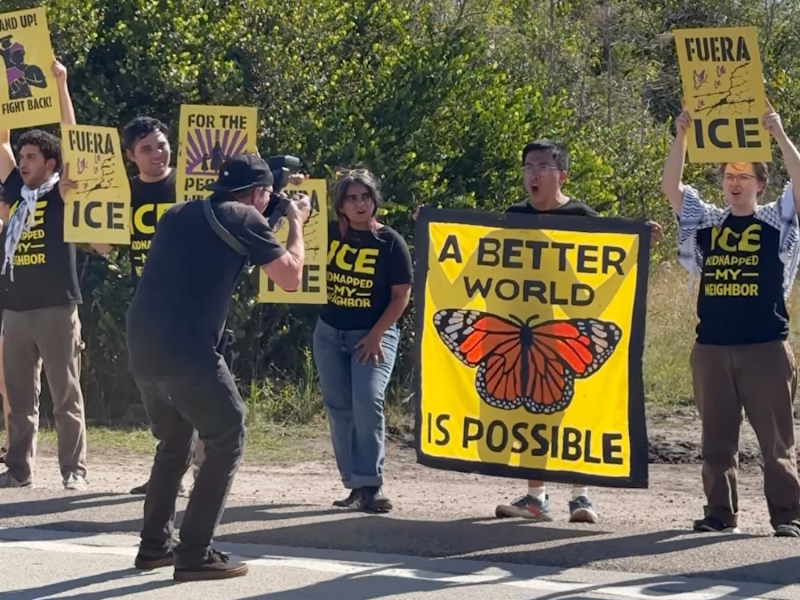 A group of protestors stands outside holding yellow and black signs. A cameraman films a man holding a large yellow banner featuring a monarch butterfly and the text, "A BETTER WORLD IS POSSIBLE."