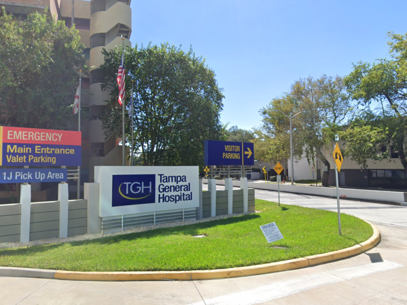 A clear, sunny photo of the Tampa General Hospital entry drive, showing multiple directional signs and the TGH logo sign set in a grassy median in front of the multi-story hospital building.