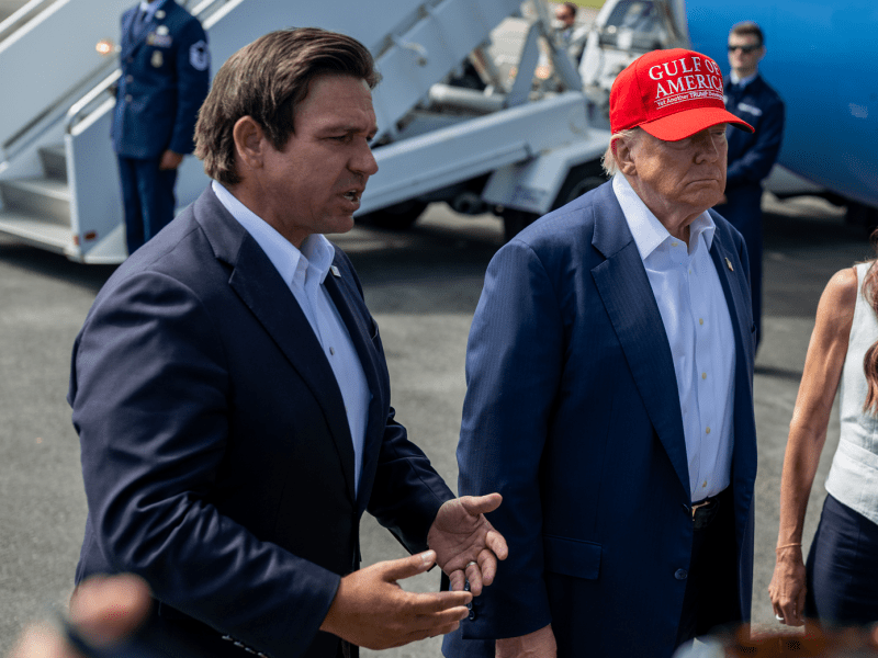 A close-up of a media event on an airfield. The central figure wears a red hat and a dark suit, flanked by two others. The person on the left is speaking with an open hand gesture, while the person on the right is looking toward the press.