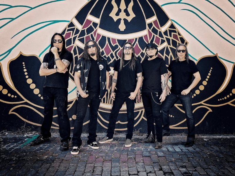 A promotional photo of the current Queensrÿche lineup, featuring Todd La Torre, Michael Wilton, Eddie Jackson, Casey Grillo, and Mike Stone, standing on a brick ground in front of a dark, stylized mural. They are all wearing black clothes and sunglasses.