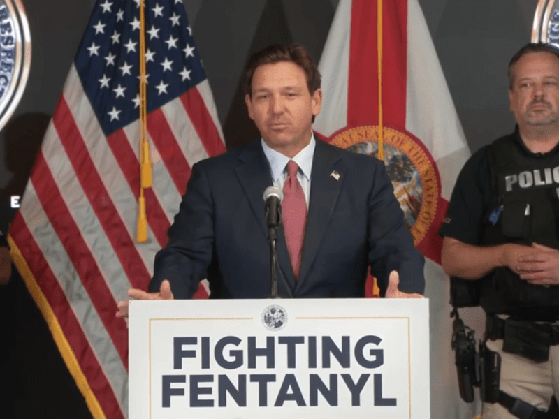 Florida Governor Ron DeSantis speaks at a podium with a sign reading "FIGHTING FENTANYL" in bold letters. He is flanked by law enforcement officers in tactical vests, with American and Florida state flags in the background, along with Florida Department of Law Enforcement insignia.