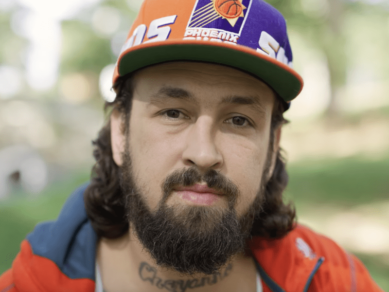 A serious portrait of a person taken in a soft focus outdoor setting, with a subtle tattoo visible on the neck. The subject is wearing a backwards baseball cap and an orange jacket over a white tank top.